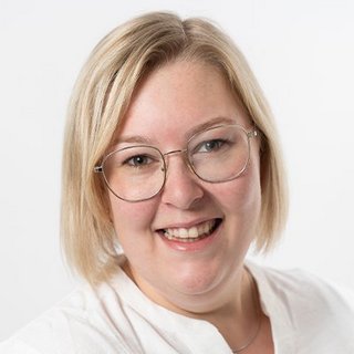 Eine lächelnde Frau mit blonden, schulterlangen Haaren und einer weißen Bluse posiert vor einem hellen Hintergrund.