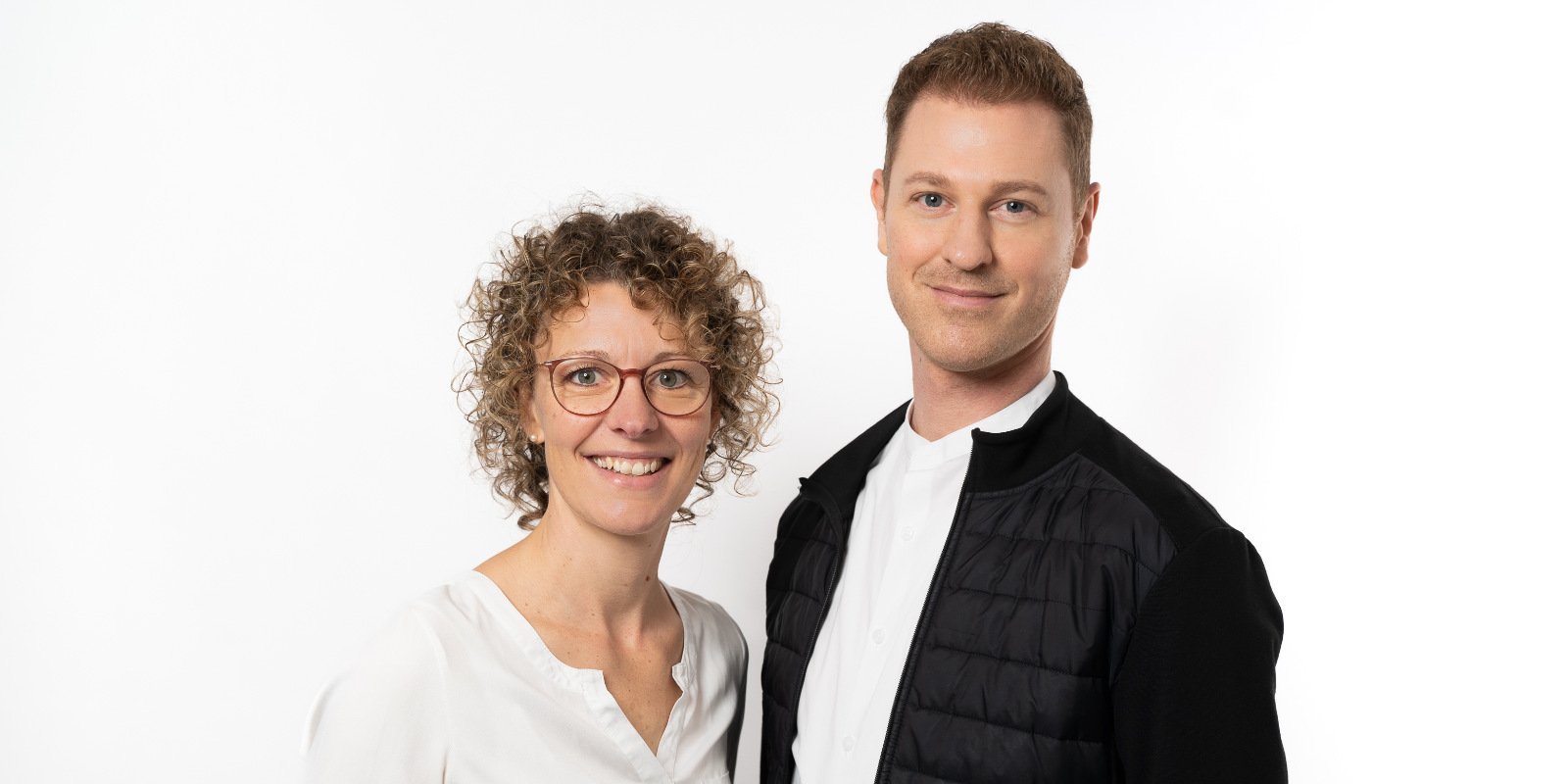 Die Frau mit lockigem Haar und Brille lächelt, während der Mann in einem schwarzen Oberteil neben ihr steht, beide strahlen positive Energie aus.