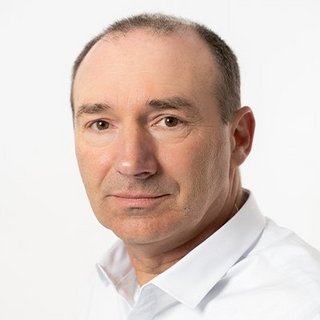 Ein Mann mittleren Alters mit kurzen, grauen Haaren und einem neutralen Gesichtsausdruck trägt ein weißes Hemd.