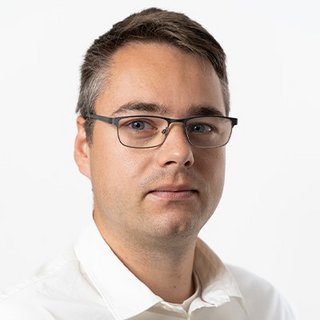 Ein Mann mit Brille und kurzem, grauem Haar schaut ernst in die Kamera, während er ein weißes Hemd trägt.