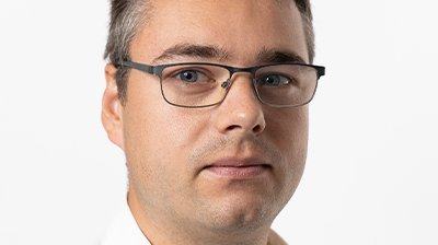 Ein Mann mit Brille und kurzem, grauem Haar schaut ernst in die Kamera, während er ein weißes Hemd trägt.