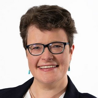 Eine lächelnde Frau mit Brille und kurzen, lockigen Haaren trägt Ohrringe und einen dunklen Blazer vor einem weißen Hintergrund.