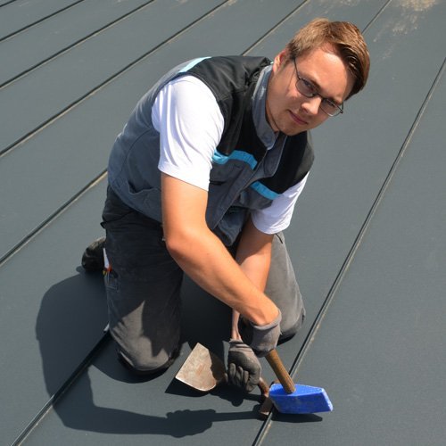 Ein Mann in Arbeitskleidung bearbeitet das Dach mit einem Hammer und einem speziellen Werkzeug, während er konzentriert aussieht.