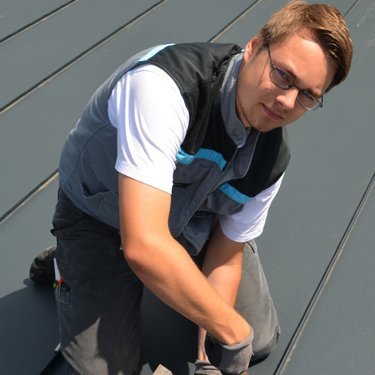 Ein Mann in Arbeitskleidung bearbeitet das Dach mit einem Hammer und einem speziellen Werkzeug, während er konzentriert aussieht.
