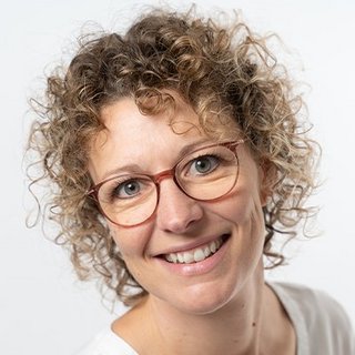 Eine lächelnde Frau mit lockigem Haar und Brille, die freundlich in die Kamera schaut, vor einem neutralen Hintergrund.