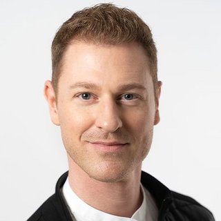 Ein Mann mit kurzem, welligem Haar und einem freundlichen Lächeln blickt direkt in die Kamera, gekleidet in einem schwarzen Oberteil.