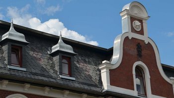 Ein detailreiches Dach mit schieferartigen Ziegeln und kunstvollen Erkern, die von weißen Wolken umrahmt werden.