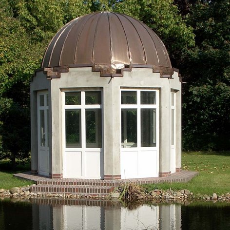 Ein elegantes Gartenhaus mit einer kupfernen Kuppel steht am Ufer eines ruhigen Teichs, umgeben von üppigem Grün.