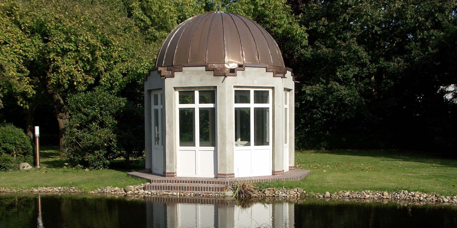 Ein elegantes Gartenhaus mit einer kupfernen Kuppel steht am Ufer eines ruhigen Teichs, umgeben von üppigem Grün.