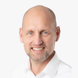 Ein lächelnder Mann mit Glatze und Bart trägt ein weißes Hemd, vor einem neutralen Hintergrund.