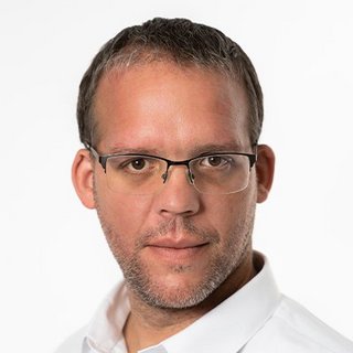 Ein Mann mit Brille und kurzem Haar schaut direkt in die Kamera, sein Gesicht zeigt einen neutralen Ausdruck.
