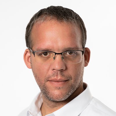 Ein Mann mit Brille und kurzem Haar schaut direkt in die Kamera, sein Gesicht zeigt einen neutralen Ausdruck.