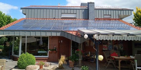 Ein modernes Haus mit einem roten Ziegeldach, das mit Solarpanelen bedeckt ist, umgeben von üppigem Grün und Blumen.